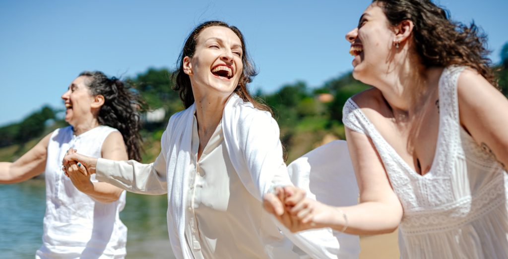 Three women joyfully holding hands and laughing on a sunny beach, wearing white outfits.
